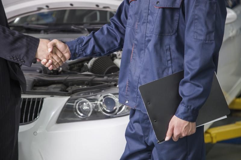 Mechanic in blue uniform shaking hands with a person in a suit near a car with open hood.