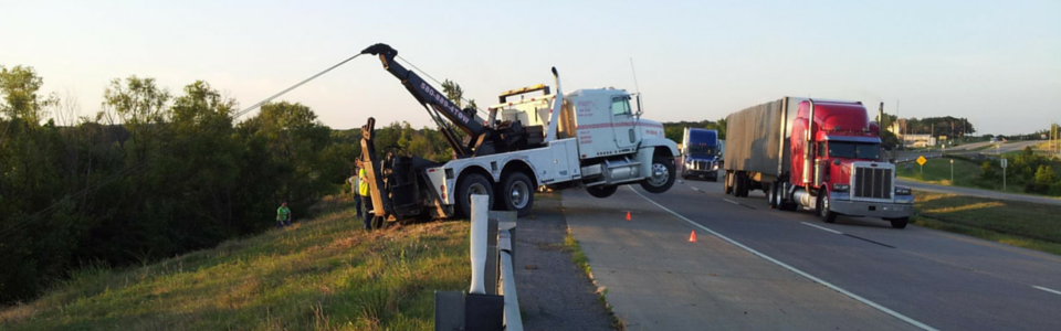 A tow truck is lifting a vehicle from a roadside ditch. Other trucks are on the highway.