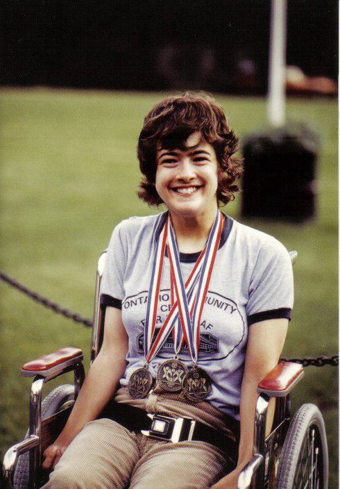 Young lady with cerebral palsy in a wheelchair smiling and wearing 3 medals of achievement around her neck