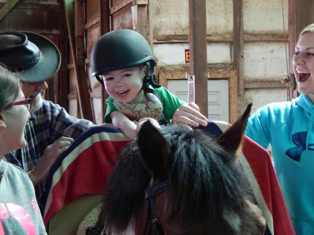 Child on top of a horse, smiling and being held up by a parent and grandparent