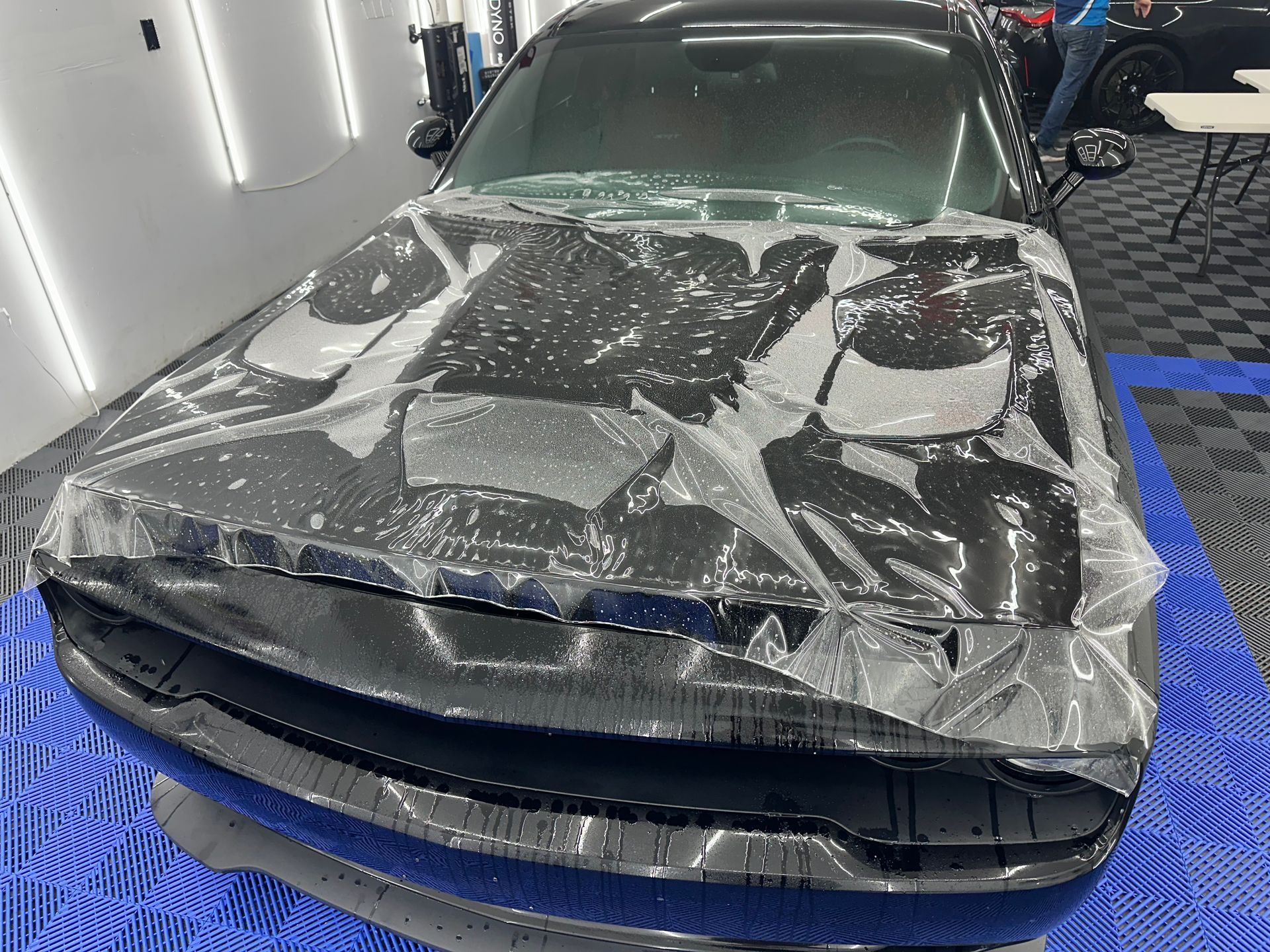 Black car hood covered in clear protective film, inside a brightly lit shop.