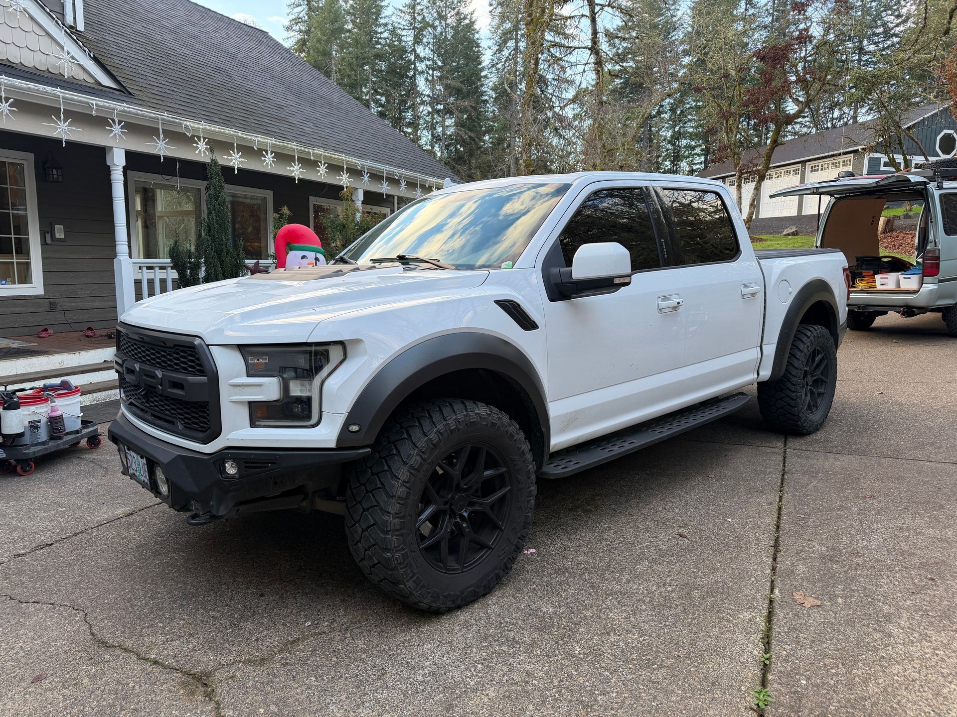 White Ford Raptor pickup truck parked on a driveway with a house in the background. Black wheels.