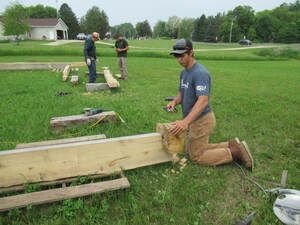 Man kneels, carving wood beam outdoors. Two others stand nearby. Green grass and buildings in background.