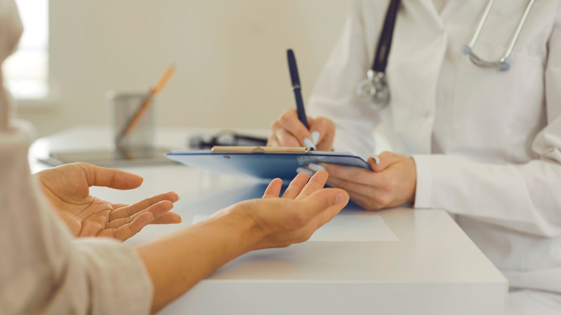 Doctor writing on clipboard, patient gesturing with open hands, in doctor's office.