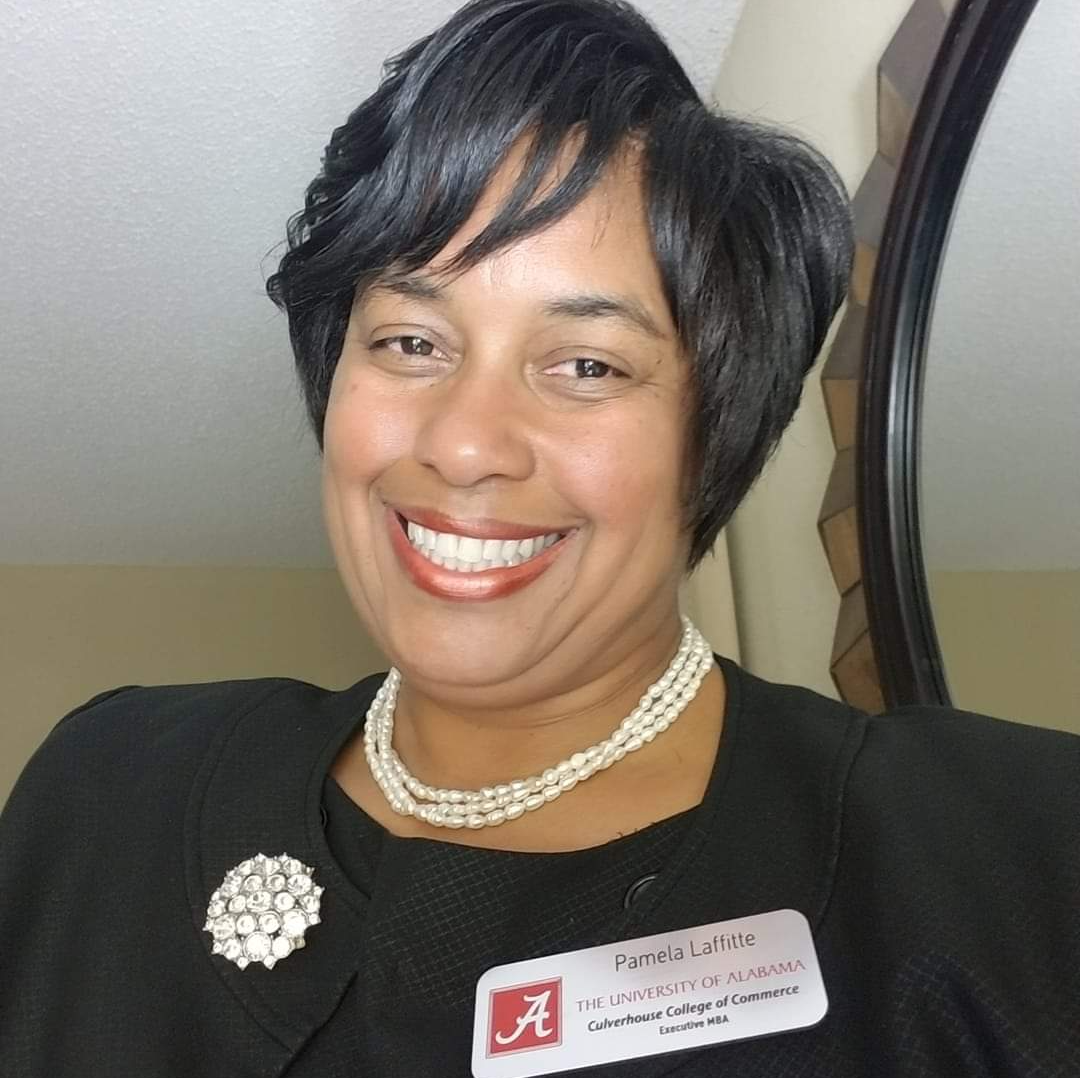 Smiling staff member in a black blazer with a name badge and pearl necklace, standing indoors.