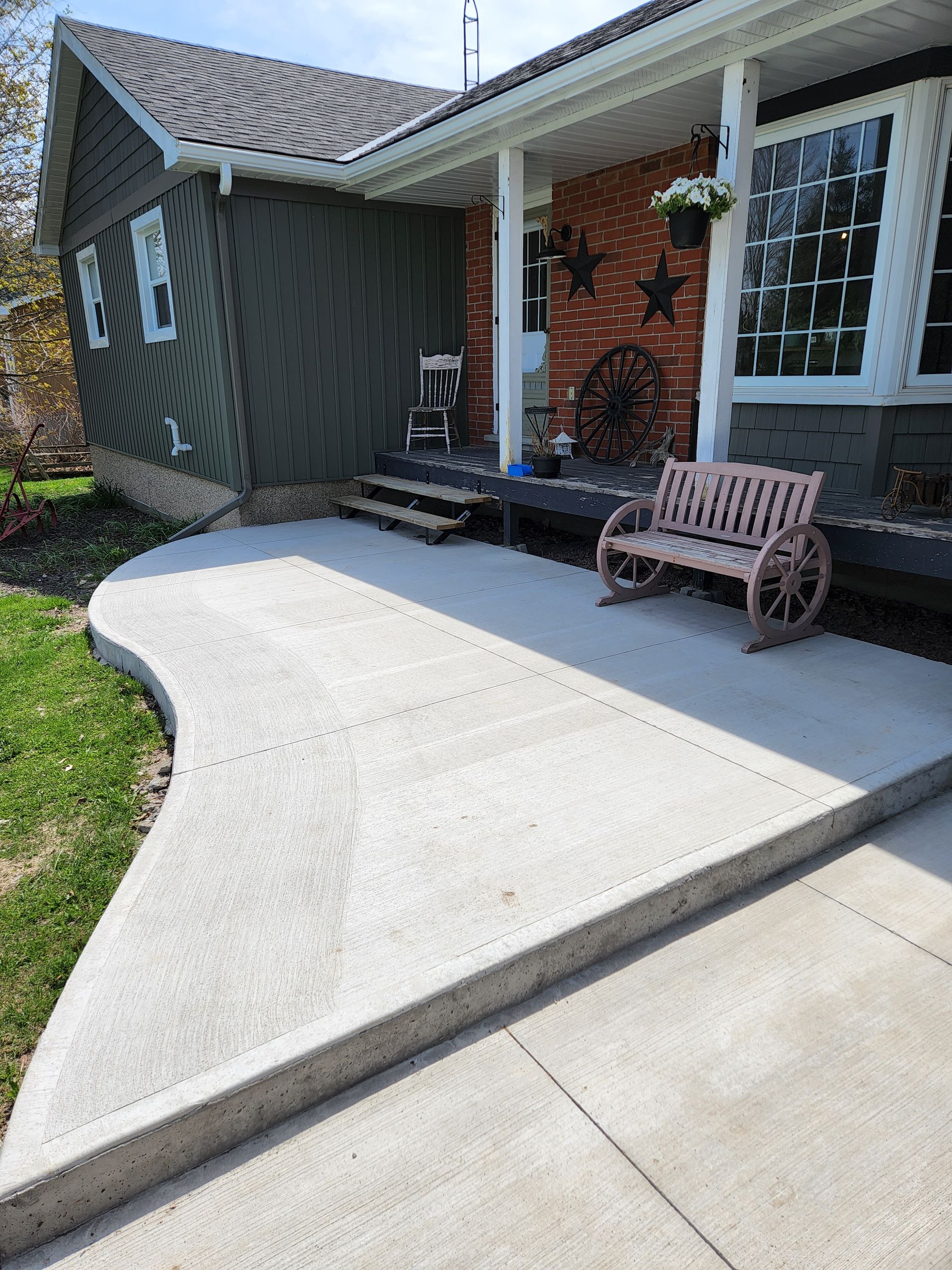 A concrete walkway leading to a house with a bench and a wagon wheel.