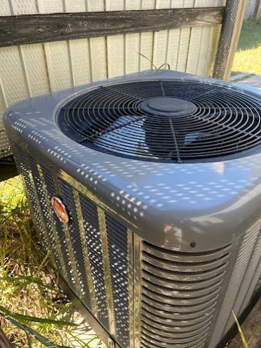 A gray air conditioner is sitting in the grass next to a fence.