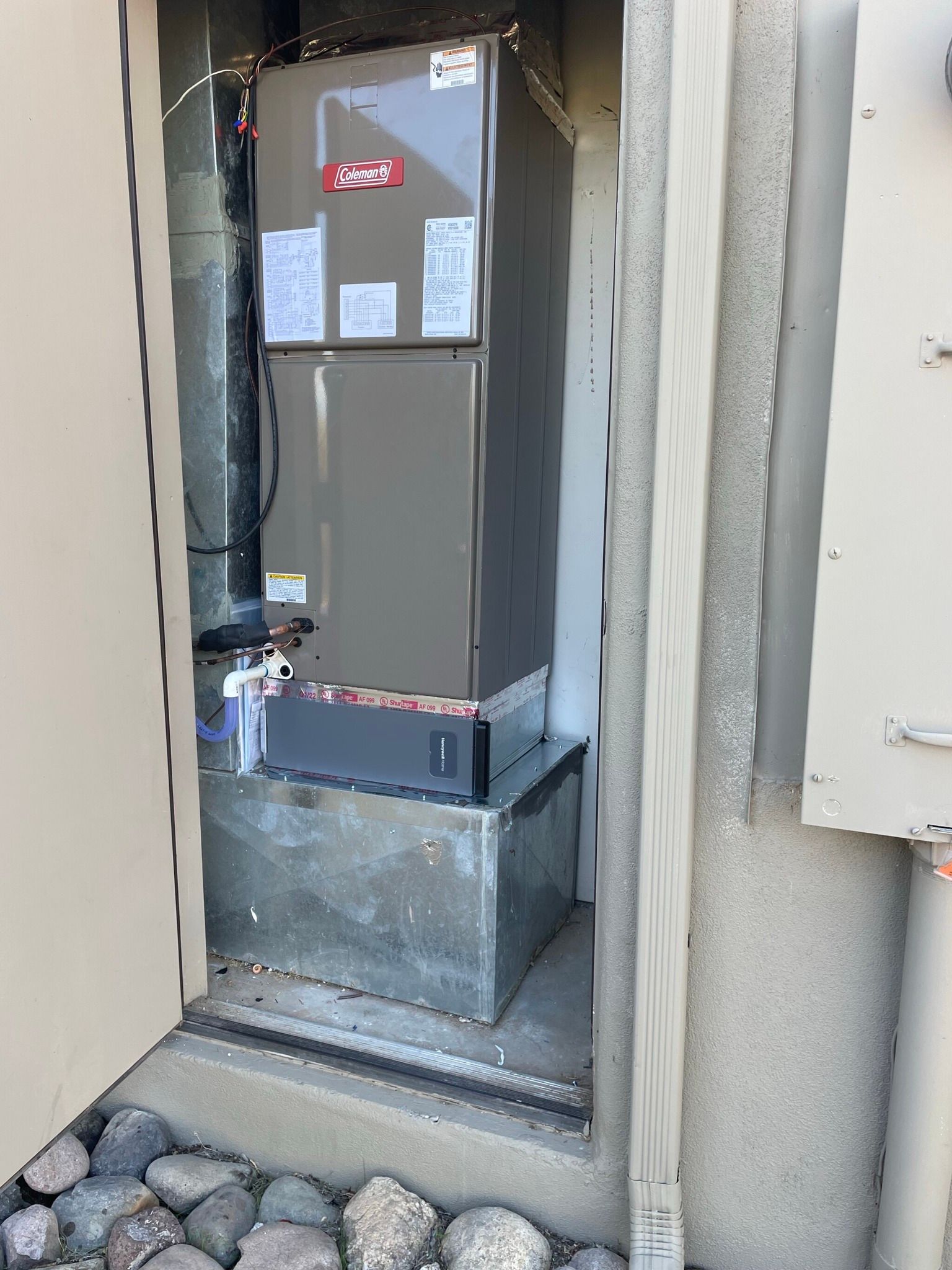 HVAC unit in a recessed outdoor alcove. Gray metallic box, ductwork, rocks, beige wall.
