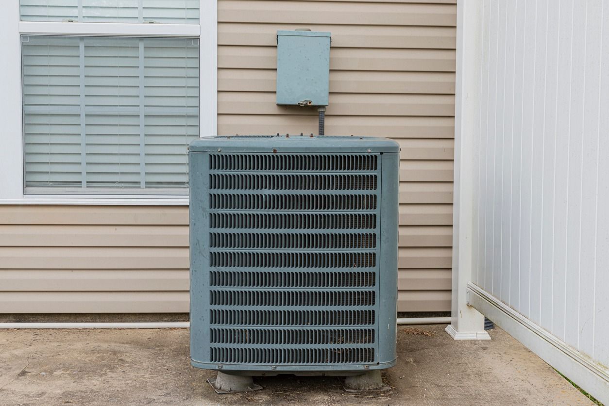 Exterior air conditioning unit against a beige-sided building, near a window and white fence.