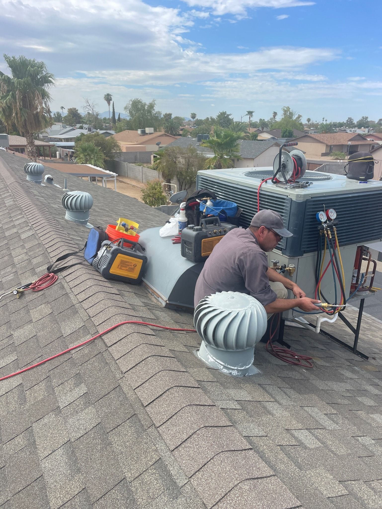 HVAC technician repairs rooftop unit, working with tools under a blue sky, surrounded by houses.