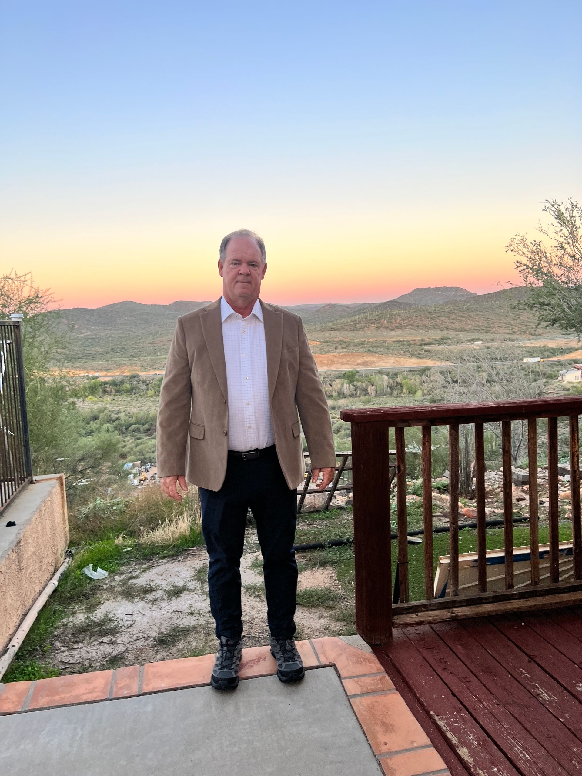Man in a tan blazer and dark pants stands on a deck with a scenic desert sunset background.