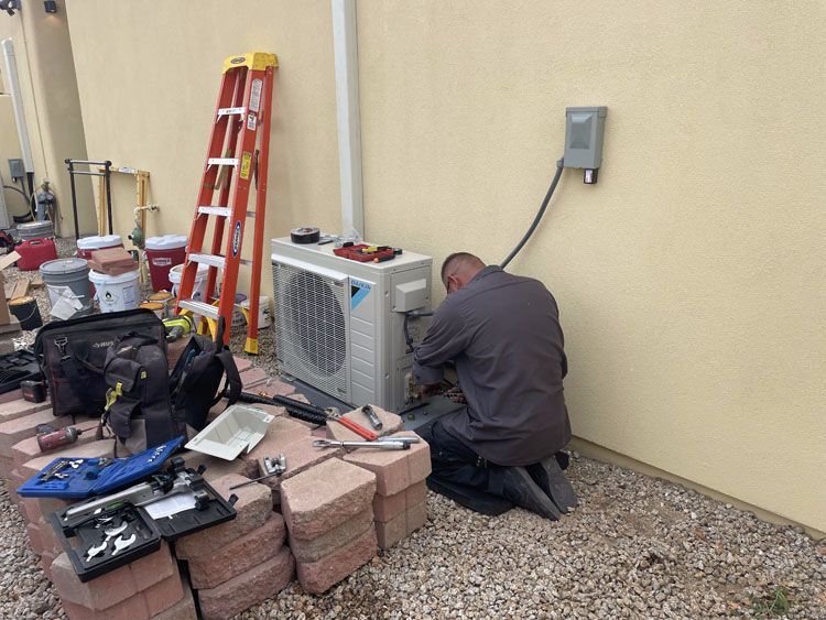 A person repairs an outdoor HVAC unit near a wall. Tools and bricks surround it.