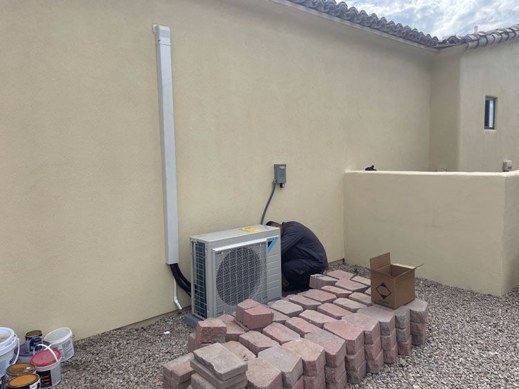 HVAC unit and electrical box mounted on tan stucco wall, surrounded by gravel and bricks.