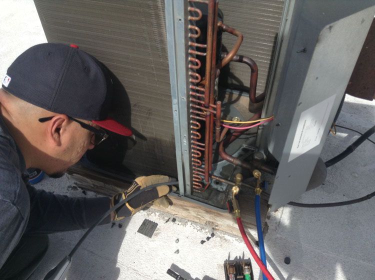 HVAC technician inspecting an outdoor unit on a rooftop. He's connecting gauges with blue/red hoses.