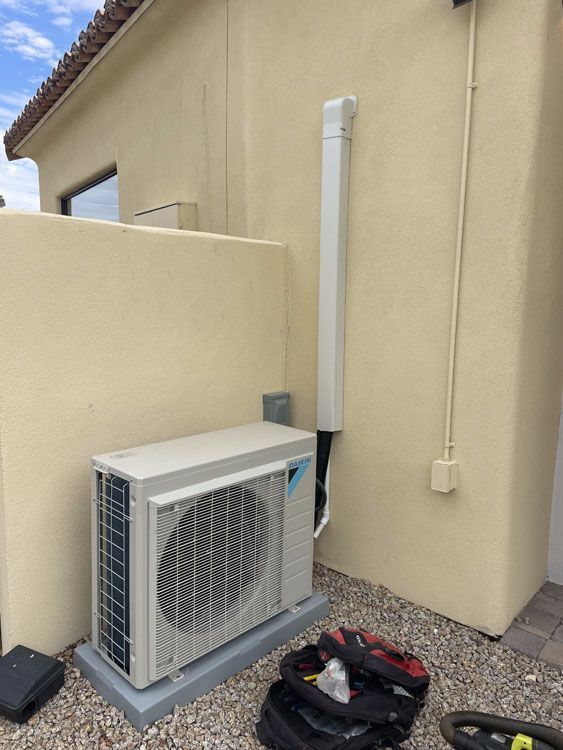 Air conditioning unit outside a stucco building, with conduit running up the wall.