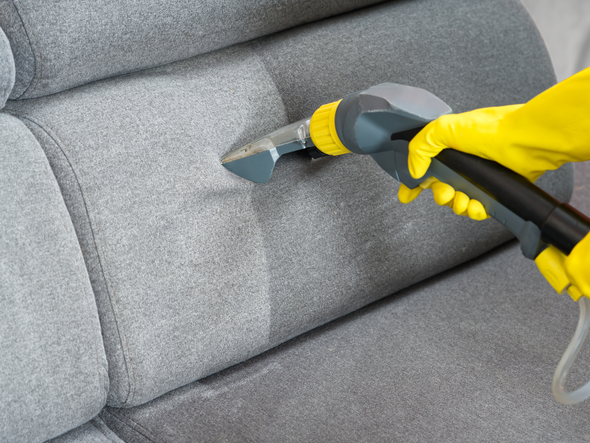 A gloved hand uses an upholstery cleaning machine to clean a gray fabric sofa, showing a clear contrast between sections.