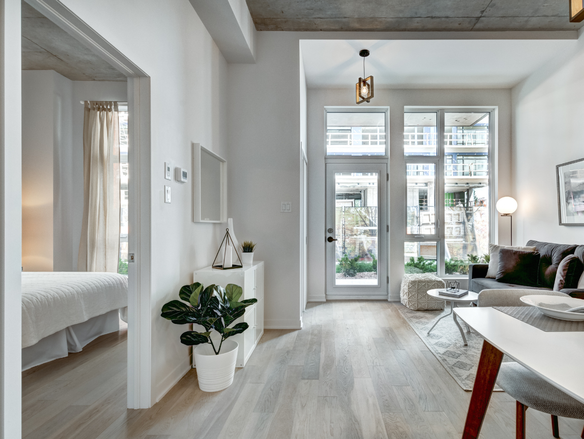 A modern, brightly lit apartment living area featuring light wood floors, a view into a bedroom, and a patio door.