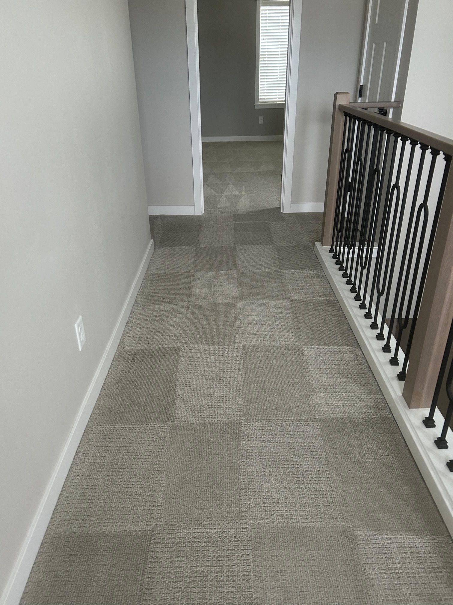 A gray carpeted hallway with a dark metal railing on the right and a doorway leading to a carpeted room in the background.