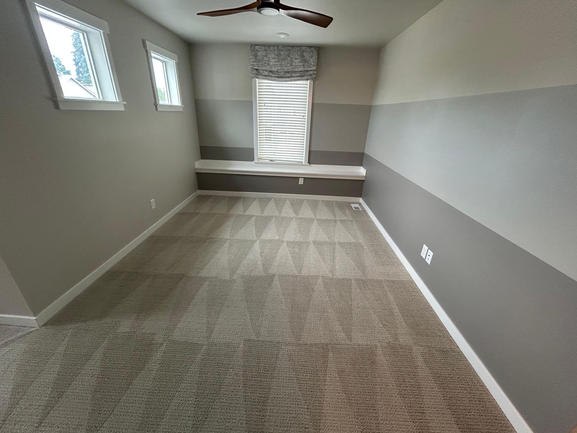 Empty room with two-tone grey walls, light carpet, ceiling fan, and a built-in window seat under a centered window.