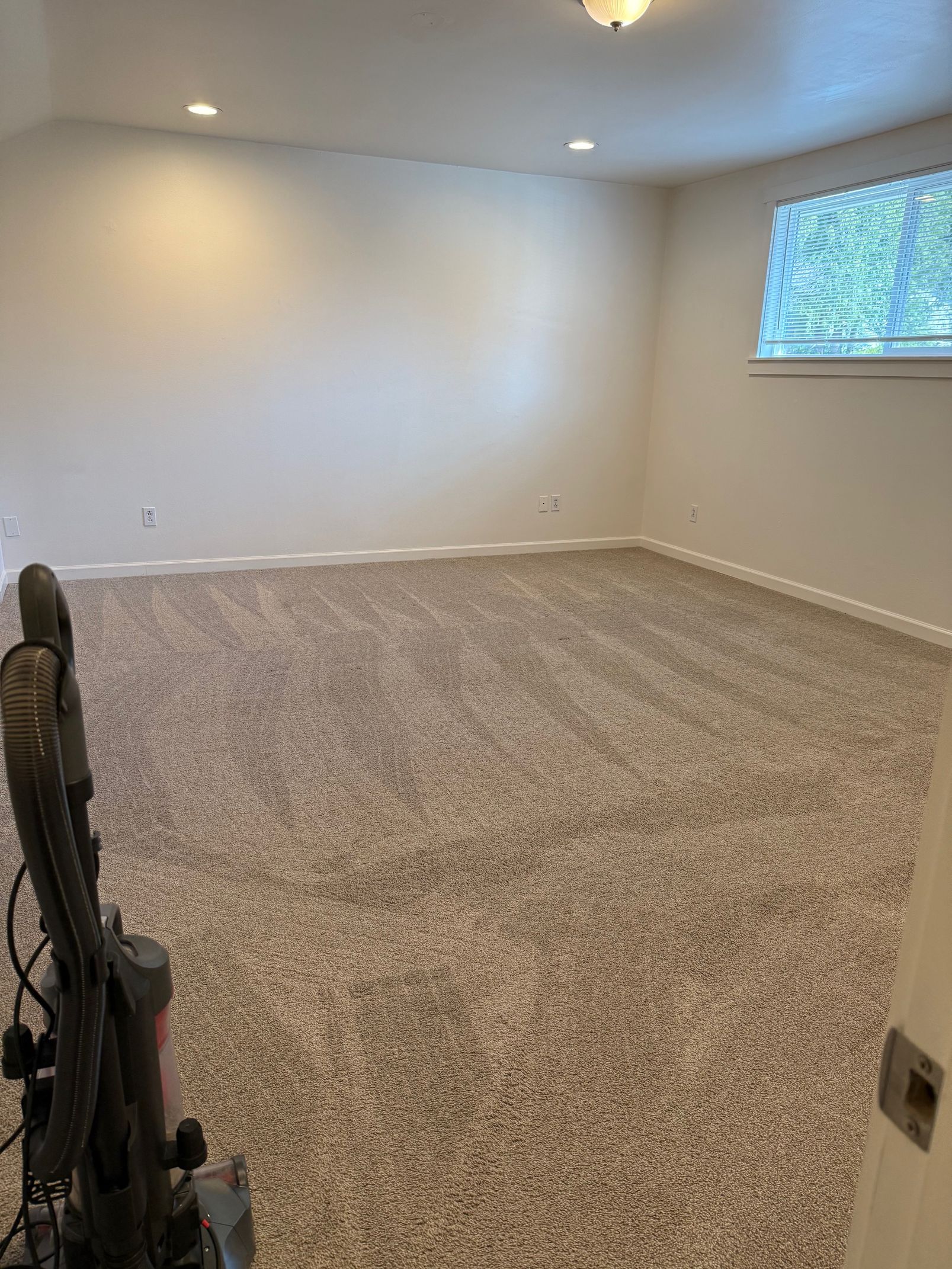 An empty carpeted room with beige walls, recessed ceiling lights, and a window with blinds on the right.