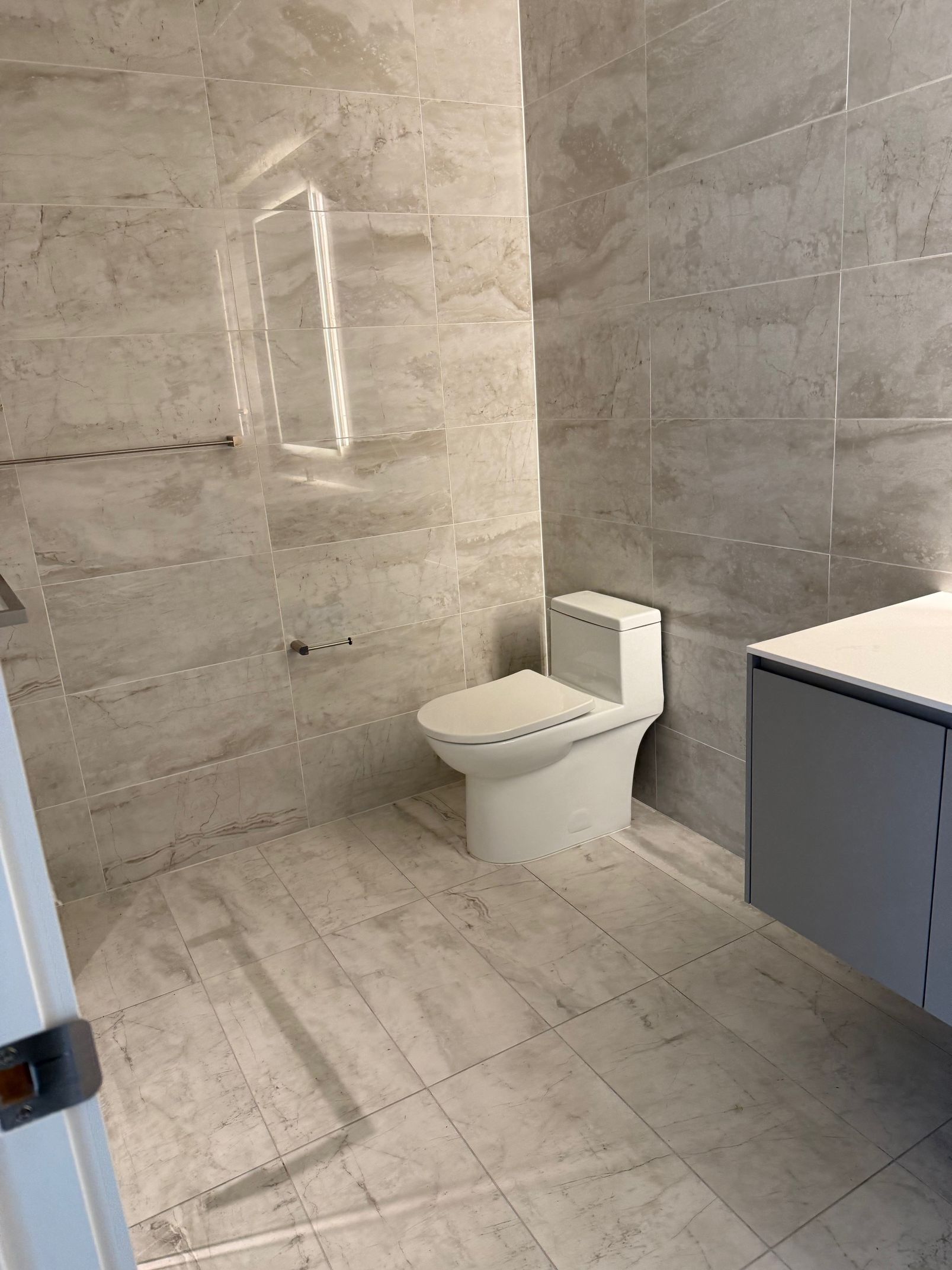 A modern bathroom featuring large-format light gray stone-look tiles, a white one-piece toilet, and a vanity cabinet.