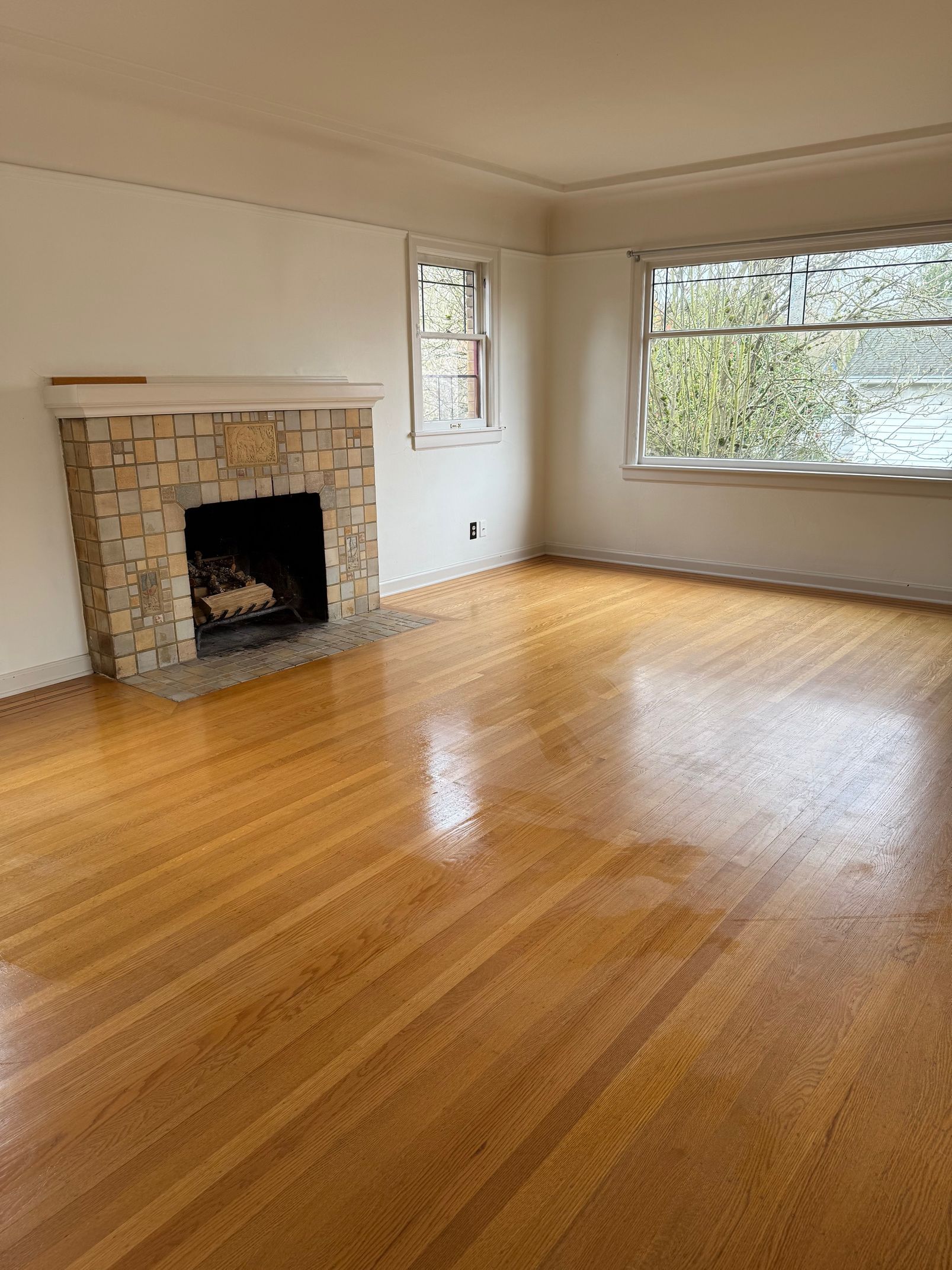 An empty living room with light-colored hardwood floors, a stone fireplace, and a large window with hanging curtains.