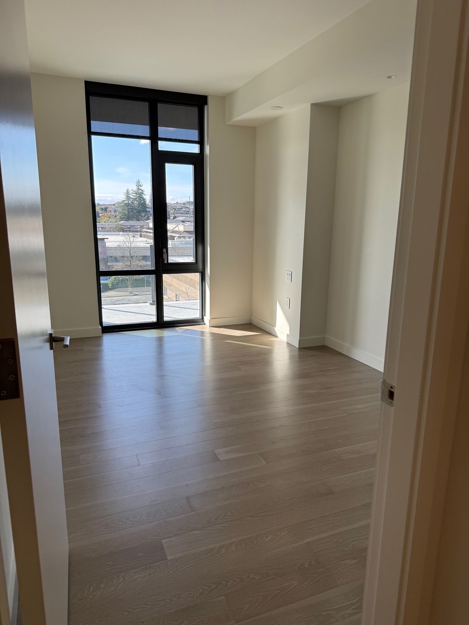 An empty, sunlit room with light wood floors, white walls, and a large floor-to-ceiling window overlooking a city street.