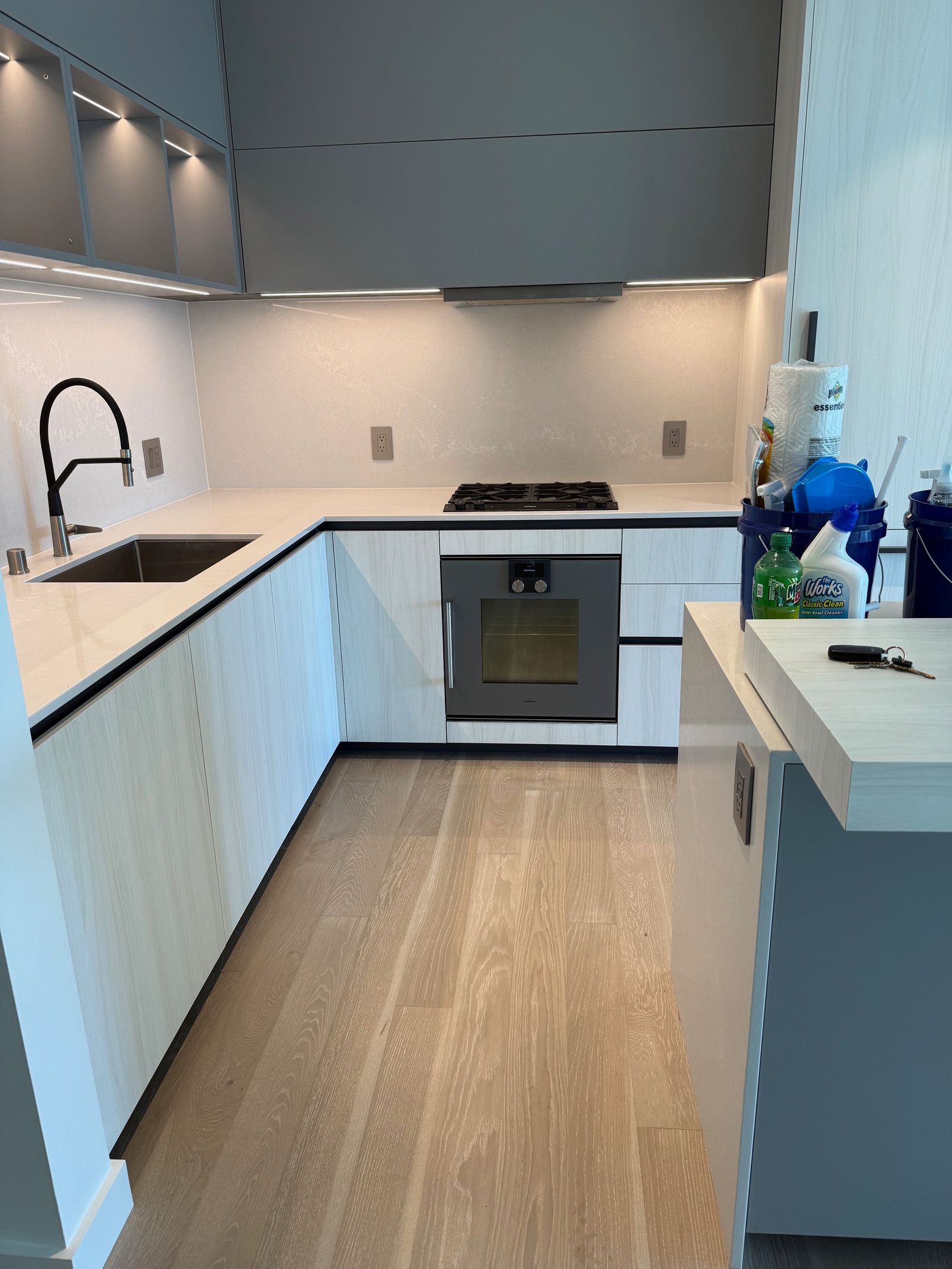 A modern kitchen featuring light wood cabinetry, white countertops, a sink, a stove, and under-cabinet lighting.