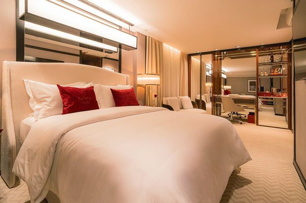 Um quarto de hotel luxuoso e bem iluminado, com uma cama macia de lençóis brancos e almofadas decorativas vermelhas, ao lado de uma sala de estar com paredes de vidro.