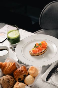Uma torrada de salmão num prato branco, um suco verde, café e uma cesta de doces variados sobre a mesa.