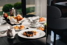 Uma mesa de café da manhã posta com um waffle coberto de chocolate, doces, um ovo cozido e café em uma sala de jantar moderna.