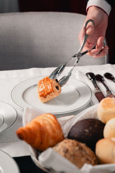 Uma mão usa uma pinça de prata para colocar um pequeno pastel em um prato branco ao lado de uma cesta de pães em uma mesa de restaurante.