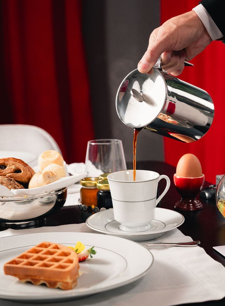 Uma mão serve café de uma jarra de prata para uma xícara de porcelana branca sobre uma mesa posta com um waffle e um ovo cozido.