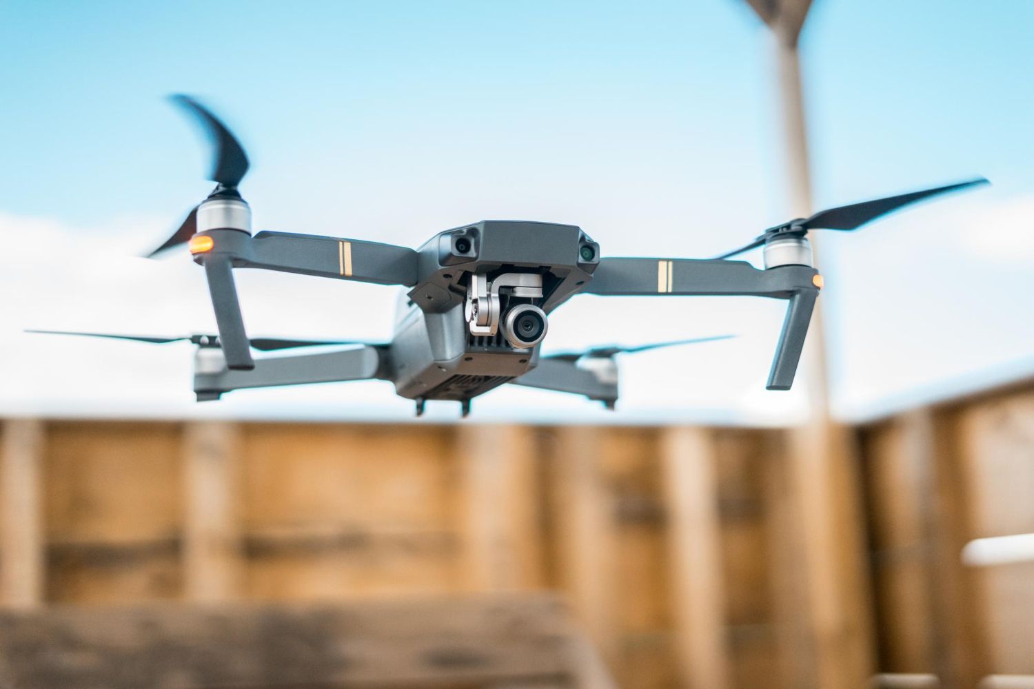 A gray drone with spinning propellers in mid-air against a blue sky and wooden backdrop.