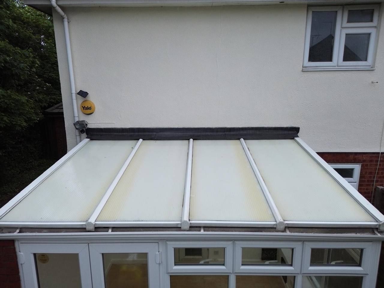 A conservatory with white frames and yellowing glass roof, attached to a cream-colored house exterior.