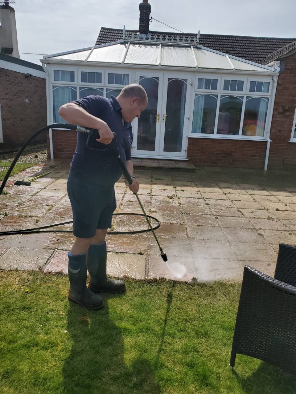 A man pressure washes a patio. He wears dark shorts, shirt, and boots, standing on grass with a sunlit patio and conservatory in the background.