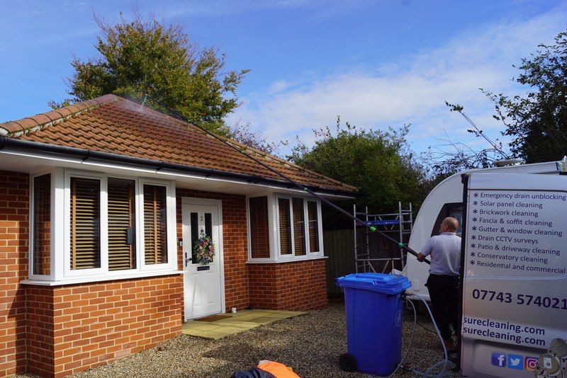 Man cleaning a bungalow roof with a long pole from a van. The house is brick with white windows and a blue bin.