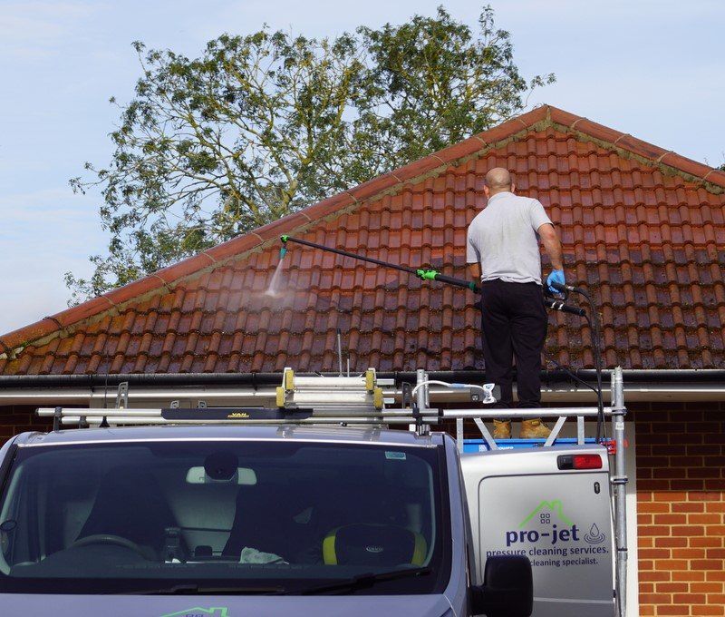 Man pressure washing a red tiled roof from the top of a van, with a tree in the background.