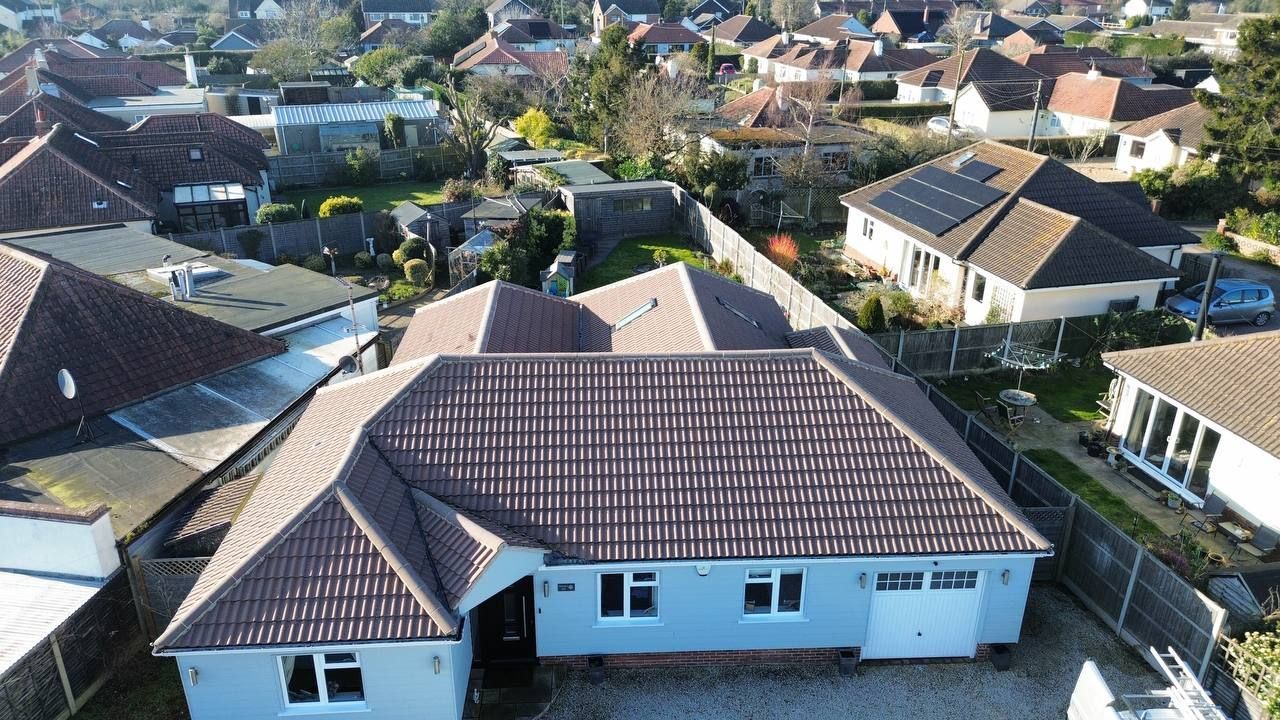 Aerial view of a light blue bungalow with a brown tiled roof, surrounded by other houses and greenery.