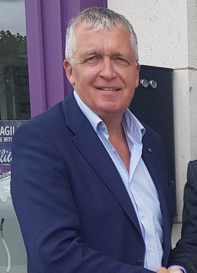 Man in blue blazer smiling, light hair, outdoors.