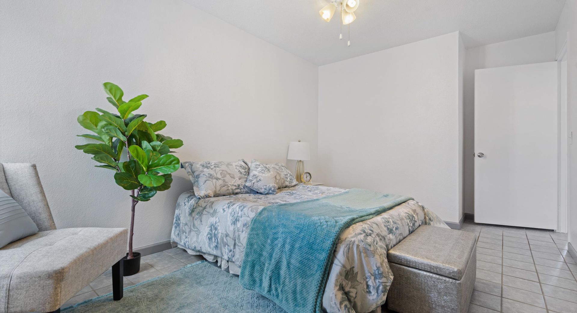 A bedroom with a bed , chair , ottoman and plant.