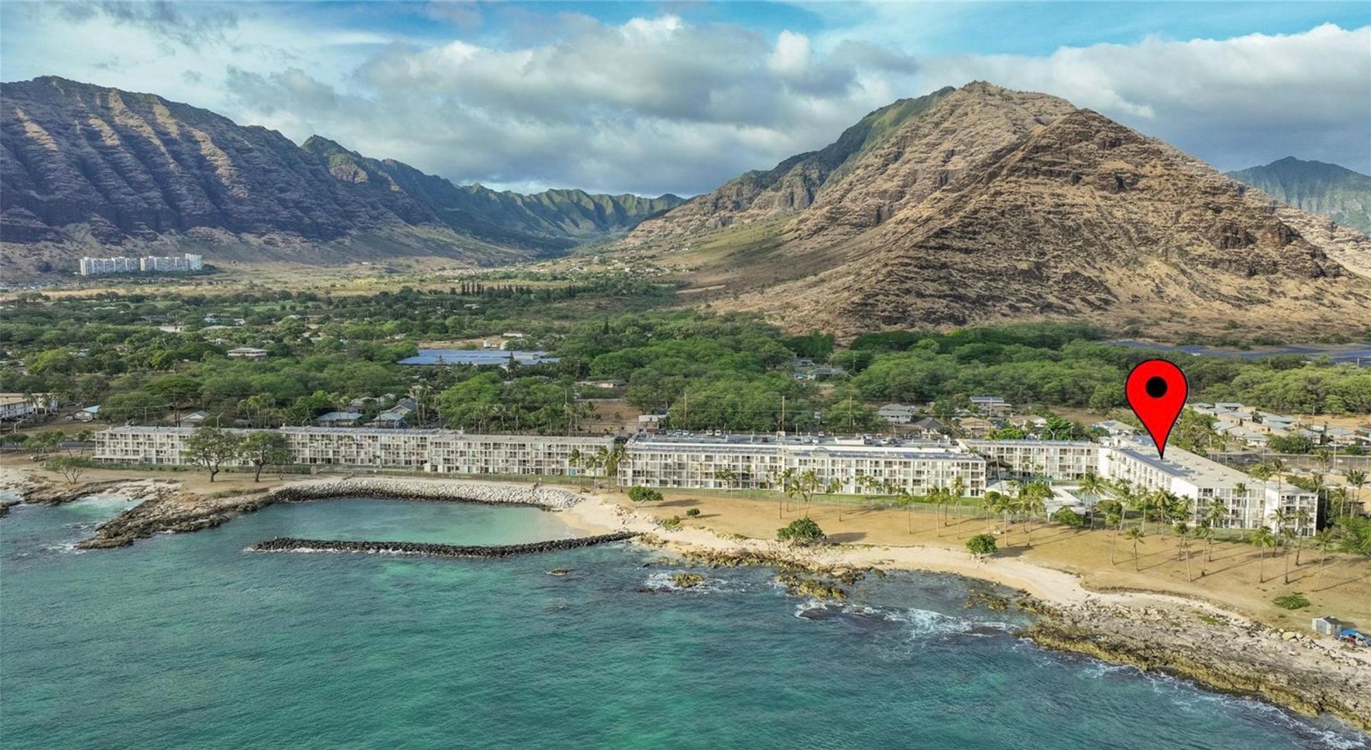 An aerial view of a beach with mountains in the background and a red pin pointing to a building.