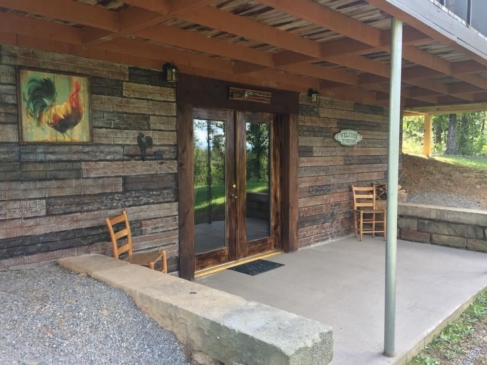 Exterior of a building with stone siding and double doors. A rooster painting is on the wall, chairs are in front.