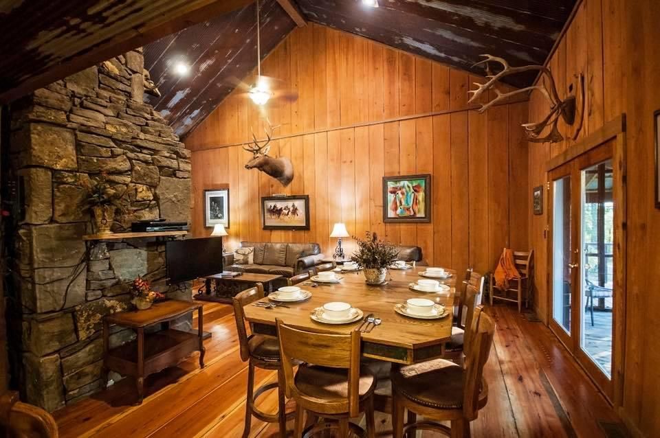 Rustic dining room with wooden walls and flooring, stone fireplace, and deer head mount.