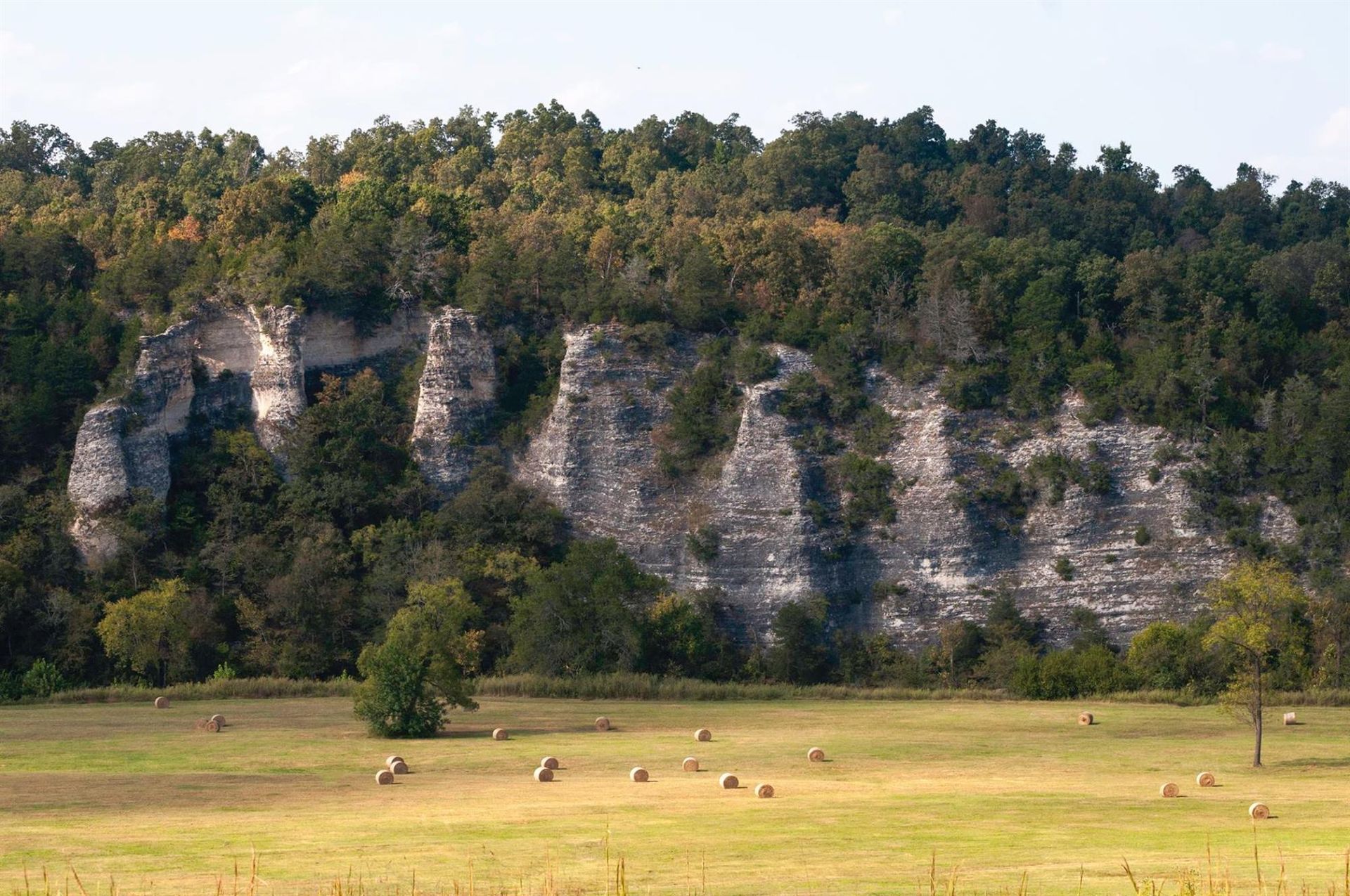Cliff face with trees at top, overlooking a field with hay bales.