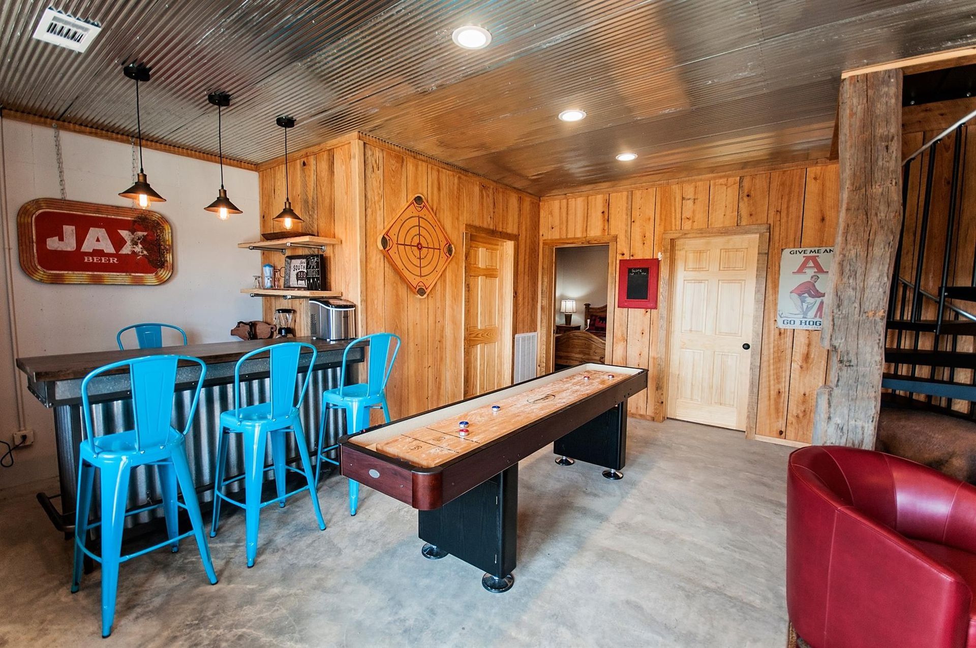 Game room with bar, shuffleboard, and blue bar stools against a wooden paneled wall with a corrugated metal ceiling.