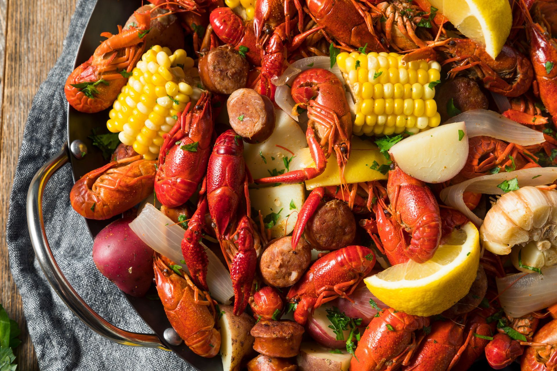 Crawfish boil with red crawfish, corn, sausage, potatoes, lemon, and onions in a metal pan.