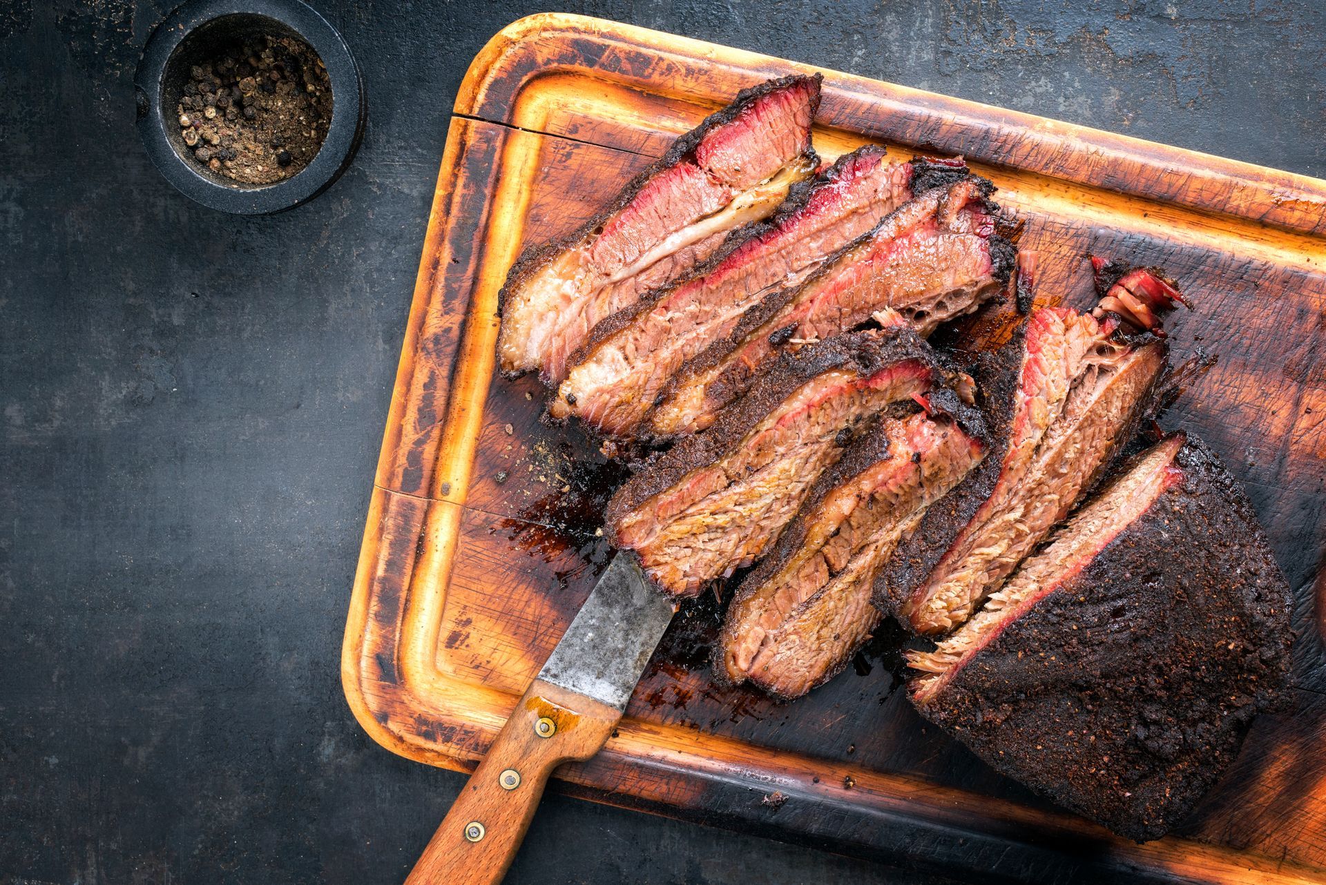 Smoked brisket sliced on a wooden cutting board with a knife and spices, top-down view.