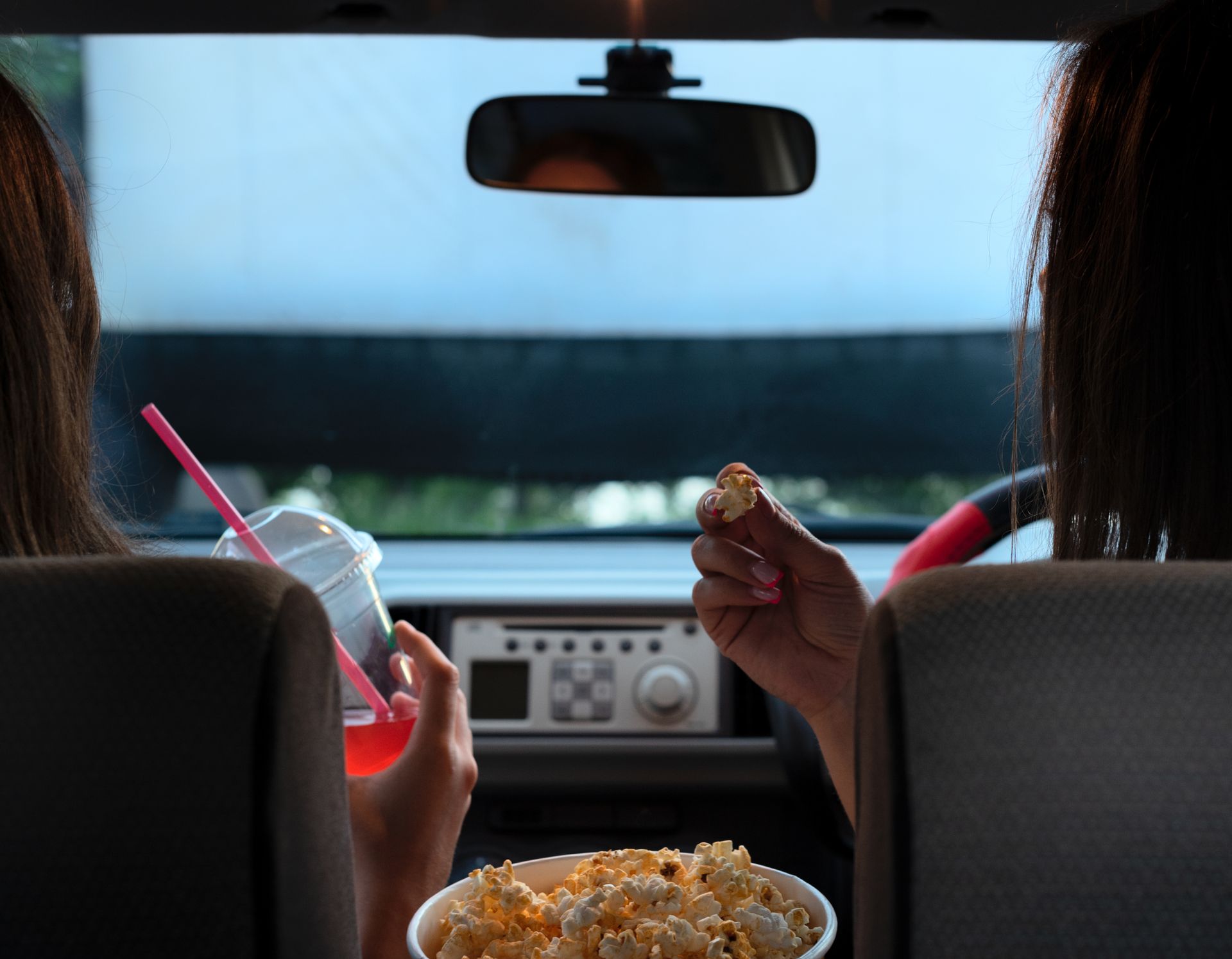 Two people in a car at a drive-in movie, eating popcorn and drinking a beverage, looking at a screen.