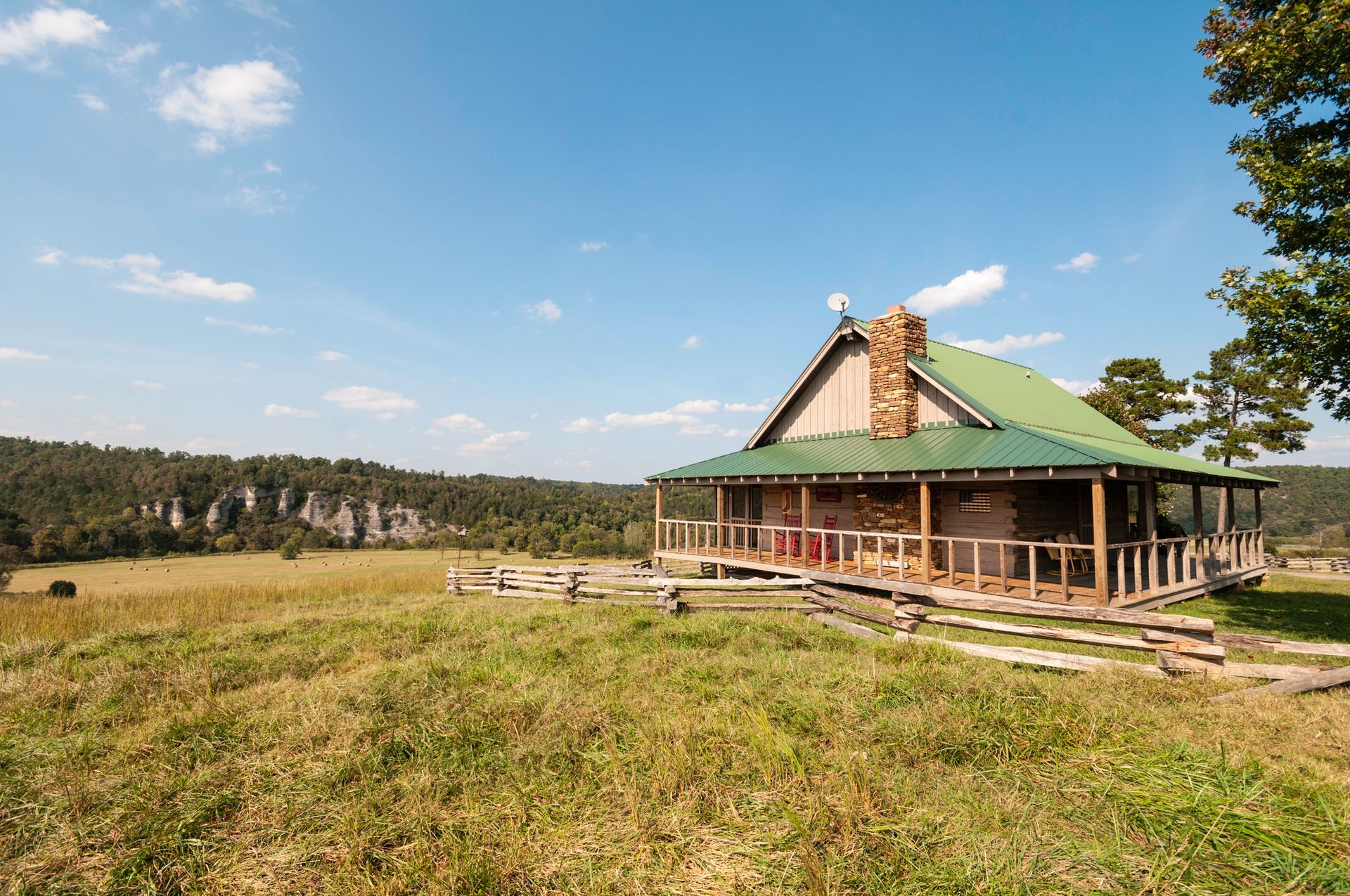 Cabin with green roof and stone chimney on a grassy hill; a wooden fence leads to the porch.